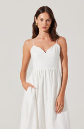 A woman is wearing a white sleeveless dress with thin spaghetti straps. The dress features a fitted bodice with a sweetheart neckline and a gathered, flowing skirt. She has her right hand in her pocket and is accessorized with large, gold hoop earrings and a gold ring on her left hand.